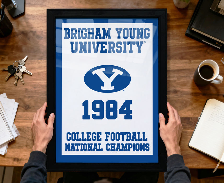 A framed poster with the text "BRIGHTAM YOUNG UNIVERSITY" and "COLLEGE FOOTBALL NATIONAL CHAMPIONS" is being held by a person's hands.