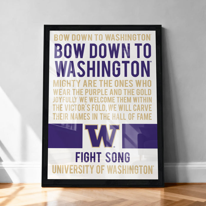 The image is a framed poster with text that reads "BOW DOWN TO WASHINGTON BOW DOWN TO WASHINGTON MIGHTY ARE THE ONES WHO WEAR THE PURPLE AND THE GOLD JOYFULLY WE WELCOME THEM WITHIN THE VICTOR'S FOLD, WE WILL CARVE THEIR NAMES IN THE HALL OF FAME". The poster is displayed on a wall, and there is a wooden floor in the background.