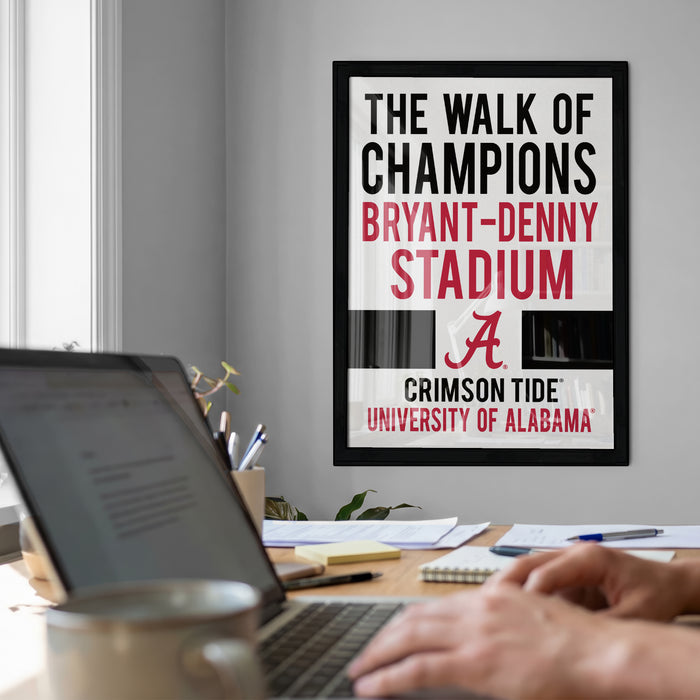 a framed poster with the text "The walk of champions" and "Bryant-Denny Stadium" on it, along with the letter "A" and the word "Crimson Tide" below. In the foreground, there is a laptop and a coffee cup on a desk, suggesting a workspace or study area.