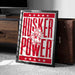 A framed poster with the text "HUSKER POWER" and a graphic of a person wearing a hat and holding a football.