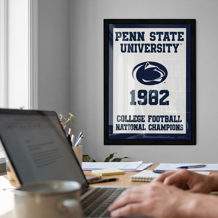 A framed poster of Penn State University from 1982 hangs on a wall, with a laptop and a coffee mug on a desk in the foreground.