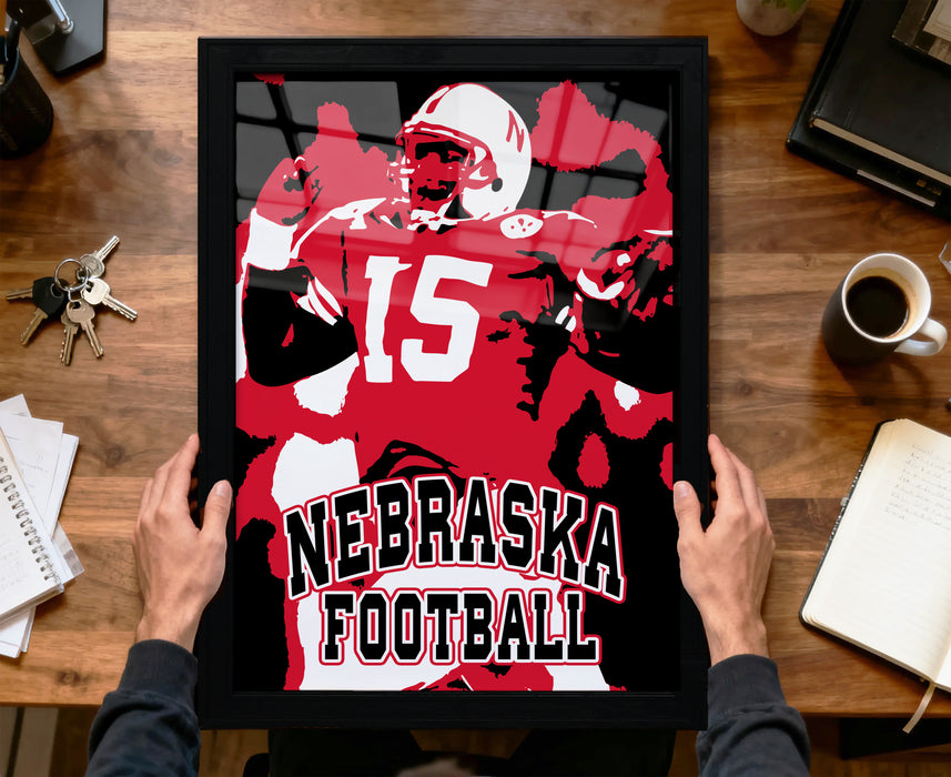 A framed poster of a football player in a Nebraska uniform is being held up by a person's hands, with a desk and various items in the background.