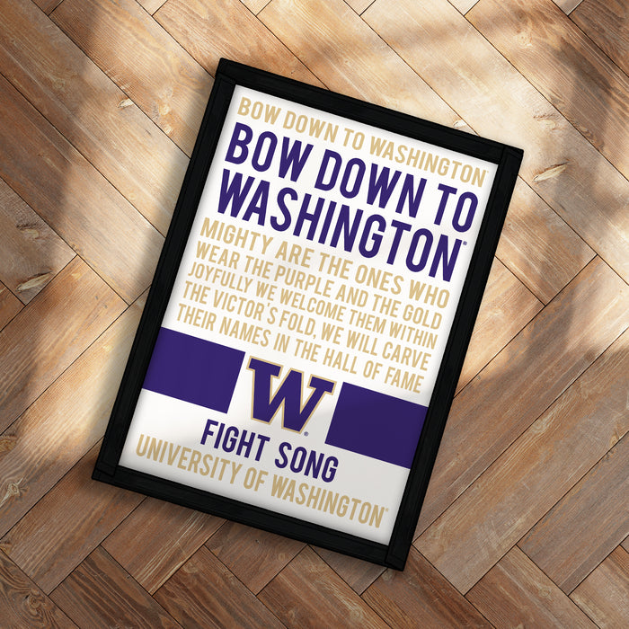 The image is a framed poster with text that reads "BOW down to Washington to WASHINGTON, MIGHTY ARE THE ONES WHO WEAR THE PURPLE AND THE GOLD JOYFULLY WE WELCOME THEM AND THE GOLD VICTOR'S FOLD WE WILL CARVE THEIR NAMES IN THE HALL OF FAME". The poster is placed on a wooden surface, and the text is in a combination of purple and white colors.