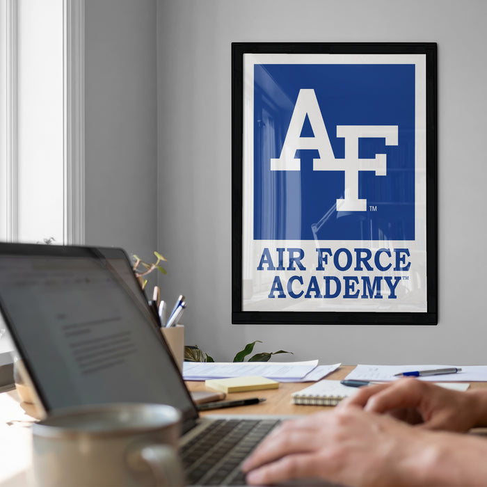 a workspace with a laptop computer, a framed Air Force Academy logo, and a person's hands typing on the keyboard.