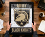 A framed poster with the text "ARMY BLACK KNIGHTS" and a graphic of a helmet and sword, held by a person's hands.