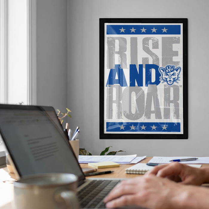 a person working on a laptop at a desk, with a framed poster featuring the text "RISE AND ROAR" in the background.