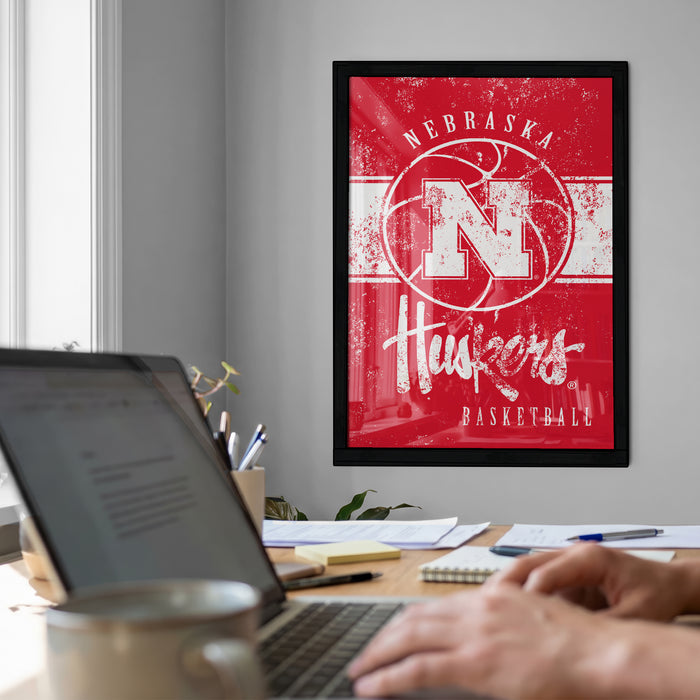 a framed poster of the Nebraska Huskers basketball team on the wall, with a person's hands typing on a laptop in the foreground.