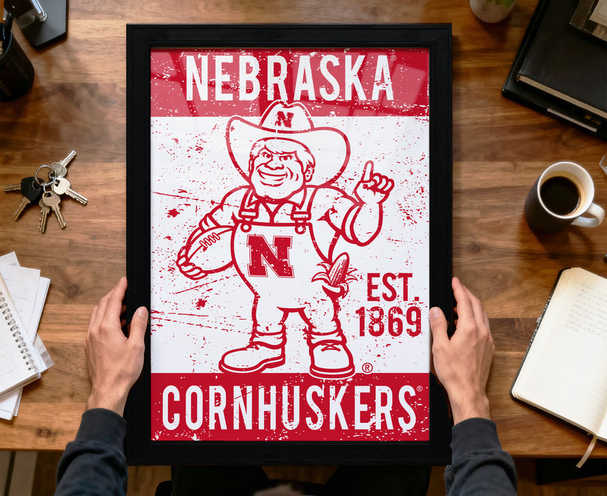 A framed poster of a cartoon character from Nebraska Cornhuskers, a sports team, is being held up by a person's hands.