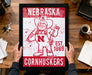 A framed poster of a cartoon character from Nebraska Cornhuskers, a sports team, is being held up by a person's hands.