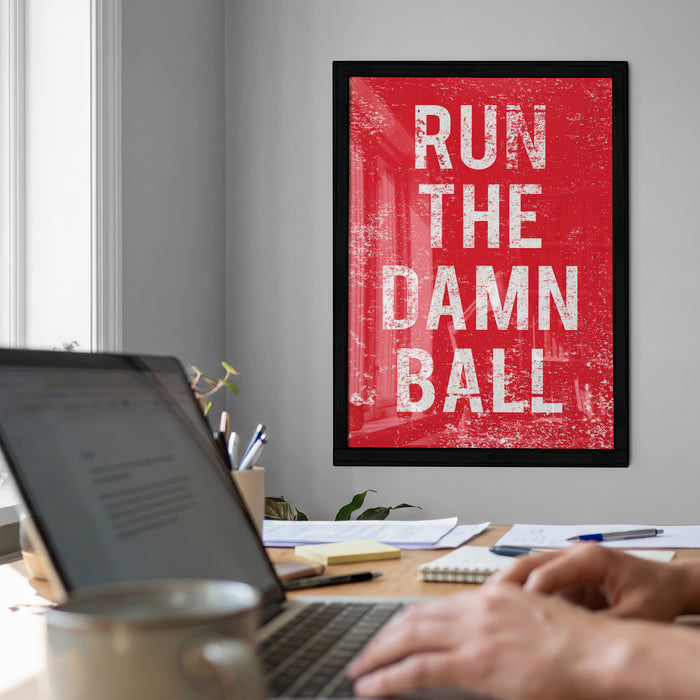 a person working on a laptop at a desk, with a framed motivational quote, "RUN THE DAMN BALL", hanging on the wall behind them.