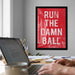 a person working on a laptop at a desk, with a framed motivational quote, "RUN THE DAMN BALL", hanging on the wall behind them.
