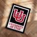 A framed poster with the University of Utah logo and text is displayed on a wooden floor.