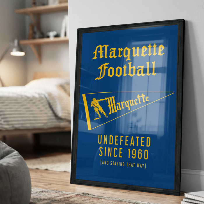 A framed poster with the text "Marquette Football" and "UNDEFEATED SINCE 1960 (AND STAYING THAT WAY)" on it, placed on a wooden floor in a bedroom.