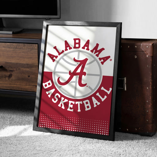 A framed poster with the Alabama basketball logo is displayed on a carpeted floor next to a wooden chest.