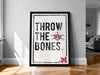 A framed poster with the text "THROW THE BONES" and a football helmet graphic, placed on a wooden floor against a white wall.