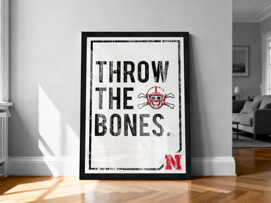 A framed poster with the text "THROW THE BONES" and a football helmet graphic, placed on a wooden floor against a white wall.