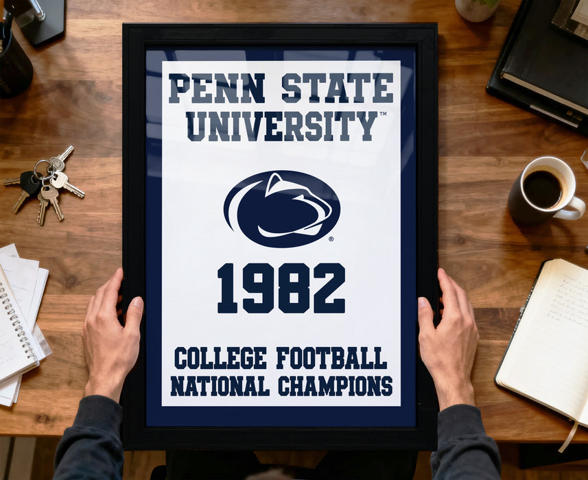 A framed poster with the text "Penn State University 1982 College Football National Champions" is being held by a person's hands, with a wooden desk and various items such as keys, a coffee cup, and a notebook in the background.