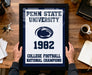 A framed poster with the text "Penn State University 1982 College Football National Champions" is being held by a person's hands, with a wooden desk and various items such as keys, a coffee cup, and a notebook in the background.