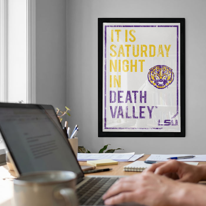 a framed poster with the text "IT IS SATURDAY NIGHT IN DEATH VALLEY" and a tiger logo, hanging on a wall above a desk with a laptop and other items.