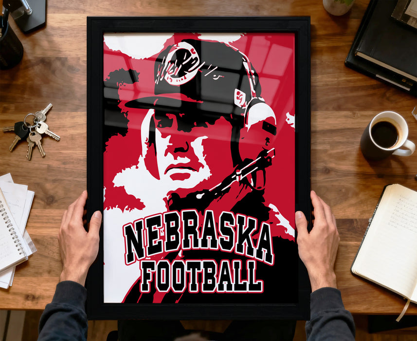 A framed poster of a football coach is being held up by a person's hands, with the text "NEBRASKA FOOTBALL" prominently displayed on the poster.