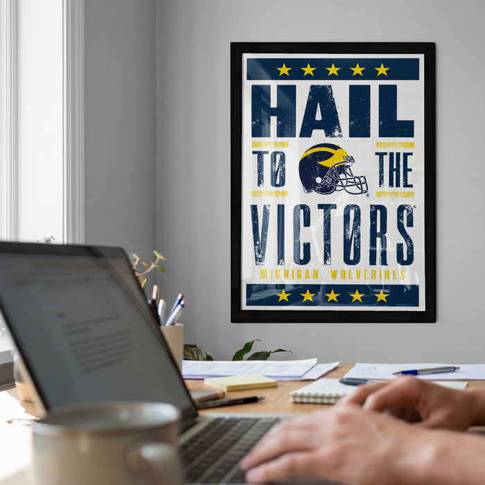 A framed poster with the text "Hail to the Victors" is displayed on a wall above a desk with a laptop and other items.