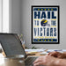 A framed poster with the text "Hail to the Victors" is displayed on a wall above a desk with a laptop and other items.