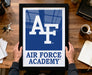A framed sign displaying the logo of the Air Force Academy is being held by a person's hands, with a desk and various items such as keys, a cup, and a notebook visible in the background.
