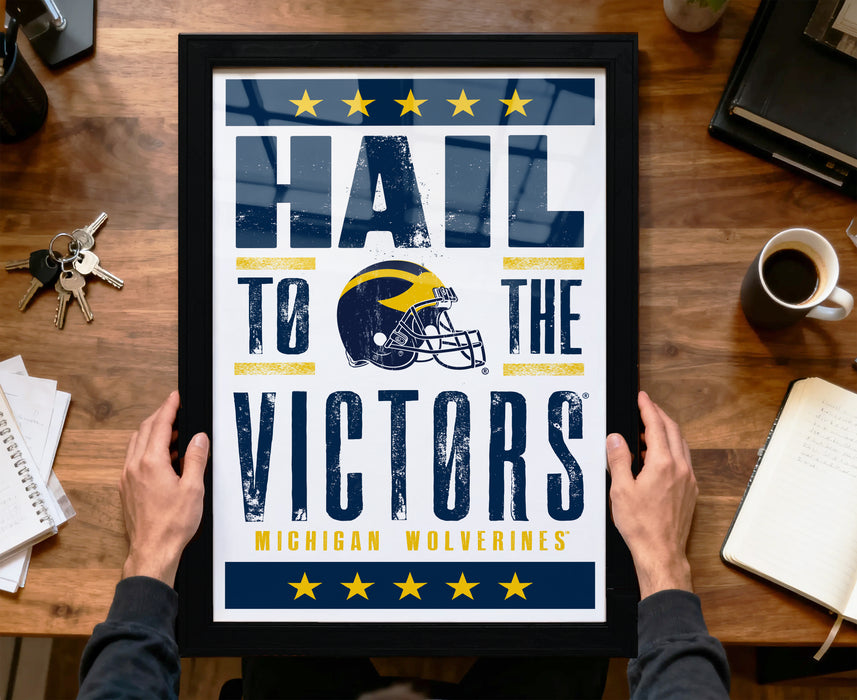 A framed poster with the text "Hail to the Victors" and a football helmet graphic, along with the text "Michigan Wolverines" is being held by a person's hands.