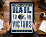 A framed poster with the text "Hail to the Victors" and a football helmet graphic, along with the text "Michigan Wolverines" is being held by a person's hands.