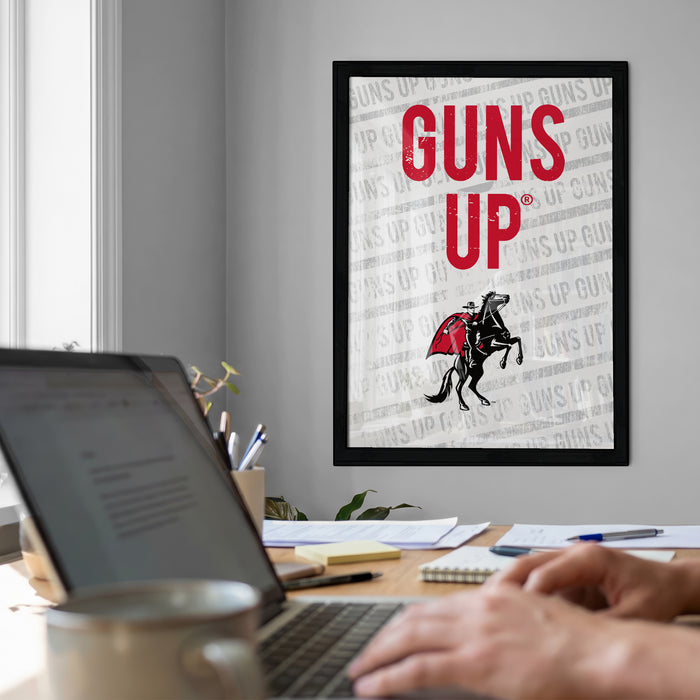 a framed poster with the text "GUNS UP" on it, which is displayed on a wall above a desk with a laptop and other items.