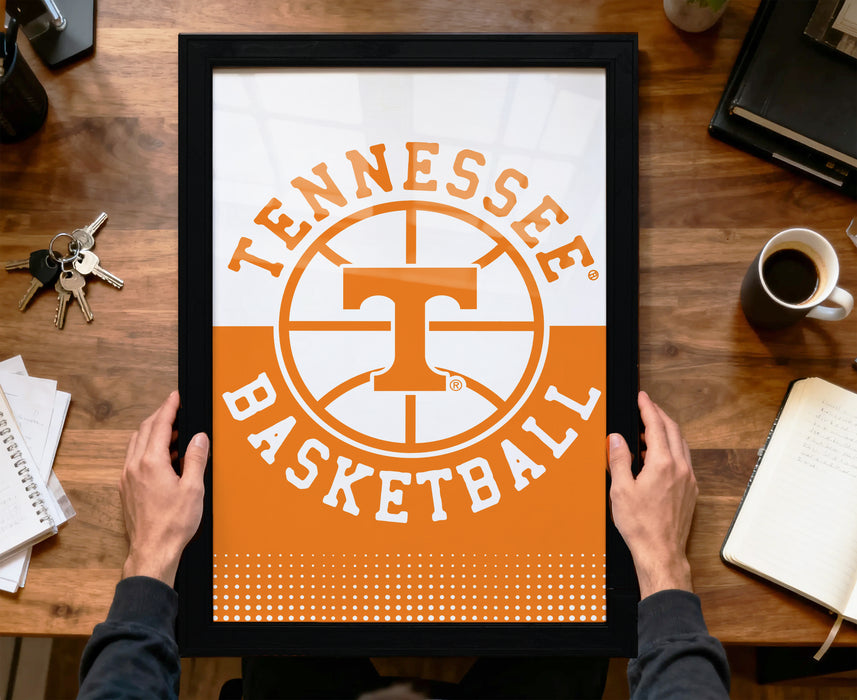 A person is holding up a framed poster with the Tennessee basketball logo on it, which is placed on a wooden desk.