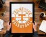 A person is holding up a framed poster with the Tennessee basketball logo on it, which is placed on a wooden desk.