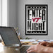 a person working on a laptop at a desk, with a framed poster on the wall behind them that reads "ENTER NIGHT".