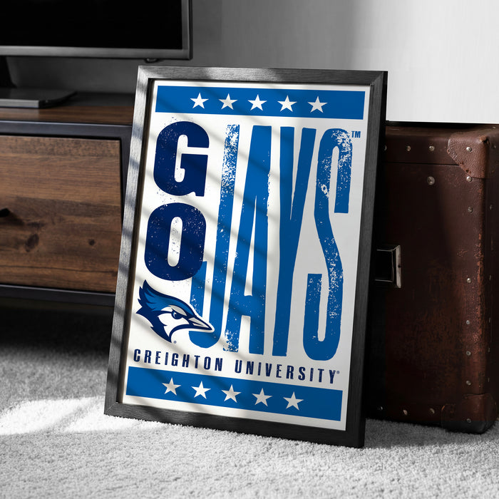 A framed poster with the text "GAYS" and a blue bird logo, placed on a carpeted floor next to a brown suitcase.