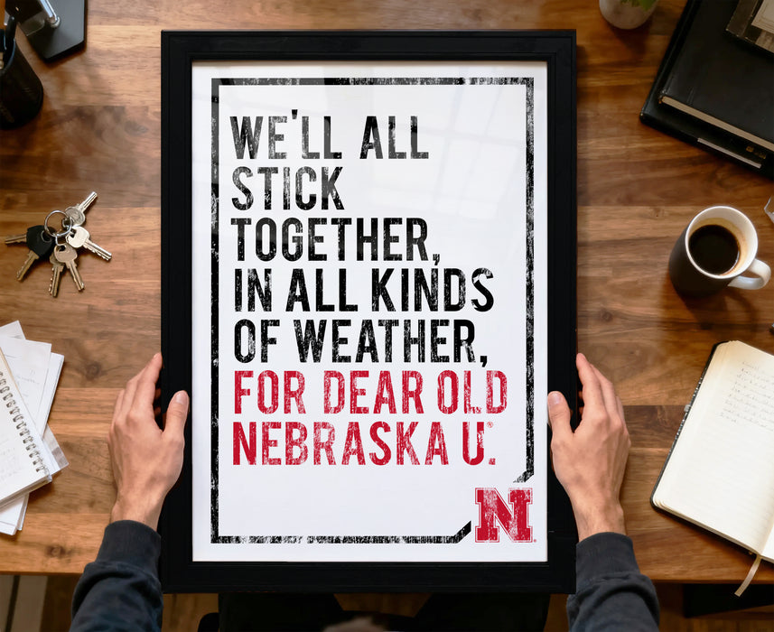 A framed poster with the text "WE'LL ALL STICK TOGETHER, IN ALL KINDS OF WEATHER, FOR DEAR OLD NEBRASKA U."