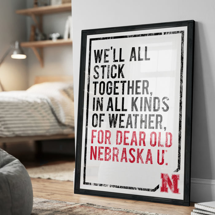 a framed poster with the text "WE'LL ALL STICK TOGETHER, IN ALL KINDS OF WEATHER, FOR DEAR OLD NEBRASKA U."
