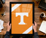 A person is holding a framed picture with the letter "T" on it, which appears to be a logo or emblem. The image is set on a wooden desk, with various items such as a cup, a notebook, and a set of keys visible.