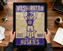 A framed poster of a mascot for Washington Huskies, a sports team, is being held up by a person's hands.