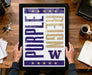 A framed poster with the text "Purple Reign" is being held by a person's hands, with a wooden desk and various office items in the background.