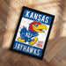 A framed poster featuring the Kansas University Jayhawks logo, a bird with a red head and yellow body, against a blue background with the text "KANSAS" and "EST. 1865" printed on it.
