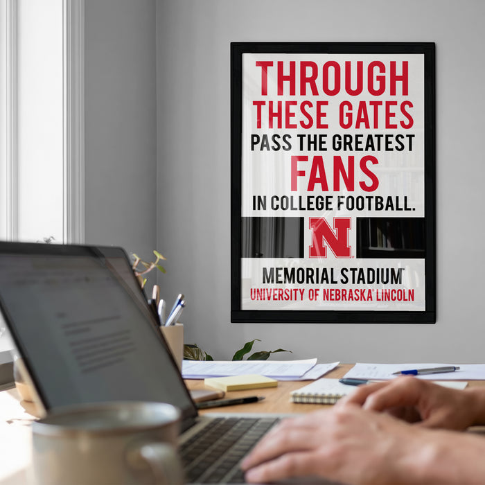 a framed poster with the text "THROUGH THESE GATES PASS THE GREATEST FANS IN COLLEGE FOOTBALL. MEMORIAL STADIUM UNIVERSITY OF NEBRASKA LINCOLN" on a wall. In the foreground, there is a laptop and a coffee cup on a desk, with a person's hands visible on the keyboard.