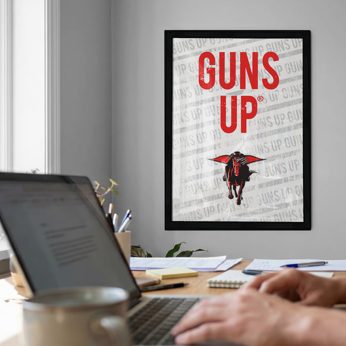 a framed poster with the text "GUNS UP" on it, which is displayed on a wall above a desk with a laptop and other items.