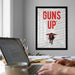 a framed poster with the text "GUNS UP" on it, which is displayed on a wall above a desk with a laptop and other items.