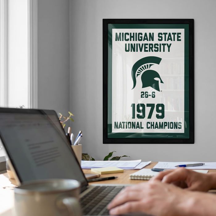 a framed poster of the Michigan State University logo hanging on a wall, with a laptop and a coffee cup on a desk in the foreground.