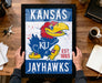 A framed poster of the Kansas University Jayhawks, a sports team, is being held by a person's hands.
