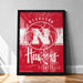 A framed poster of the Nebraska Huskies basketball team, featuring the team's logo and name on a red background.