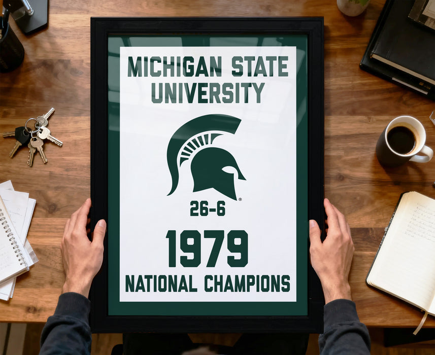 A framed poster with the Michigan State University logo and text, held by a person's hands, celebrating the university's 26-6 victory in the 1979 National Champions.