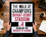 A framed sign with the text "THE WALK OF CHAMPIONS BRYANT-DENNY STADIUM A" is being held by a person's hands.