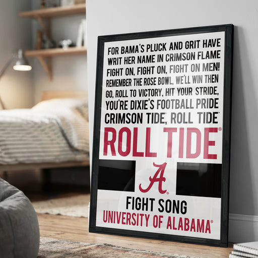 a framed poster with text that reads "For Bama's Pluck and Grit Have WRIT HER NAME IN CRIMSON FLAME FIGHT ON, FIGHT ON, FIGHT ON MEN! REMEMBER THE ROSE BOWL, WELL WIN THEN GO, ROLL TO VICTORY, HIT YOUR STRIDE, YOU'RE DIXIE'S FOOTBALL PRIDE CRIMSON TIDE, ROLL TIDE". The poster is displayed on a wall, and there is a bed and a lamp visible in the background.