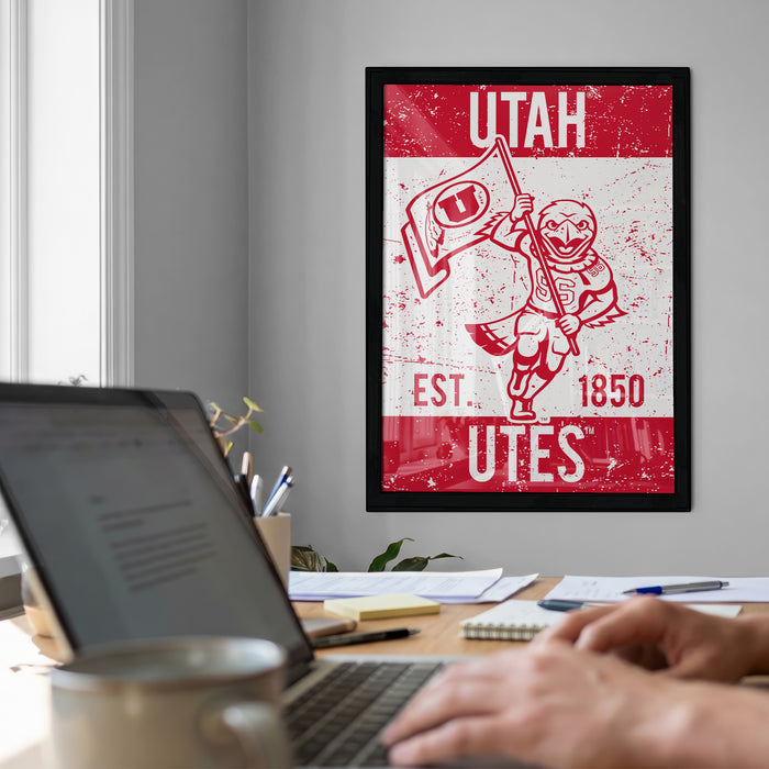 a framed poster of a red and white design with a person holding a flag, along with the text "UTAH EST. 1850 UTES". The poster is hanging on a wall, and there is a laptop and a coffee cup on a desk in the foreground.